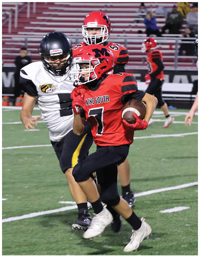 A person in a red football uniform with a helmet is running with a football while being pursued by people in black and white uniforms on a football field. Description generated by AI