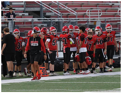 A group of people wearing red and black football uniforms and helmets stand on the sidelines of a football field with empty bleachers in the background. Description generated by AI