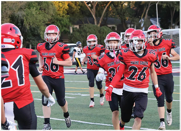 Several people wearing red football uniforms with helmets are running on a sports field with a goalpost and trees in the background. Description generated by AI