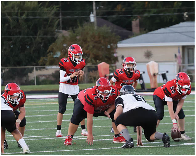 Several people wearing red and black football uniforms are positioned on a field with one person in a white and black uniform crouched in front of them, preparing for a play. Description generated by AI