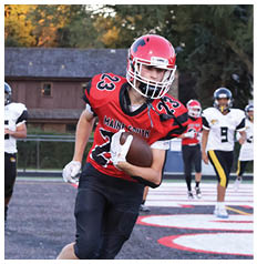 Three people wearing football uniforms are on a field with two running toward the camera and one running away, with trees and a building in the background. Description generated by AI