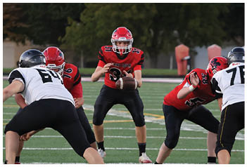 People in football uniforms are positioned on a field with one person holding a football ready to pass while others prepare to block or receive. Description generated by AI