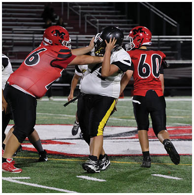 Two people in red and black football uniforms are blocking a person in a white and black uniform on a football field during a game. Description generated by AI