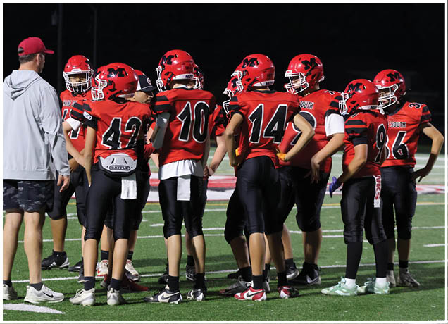 A group of people wearing red and black football uniforms and helmets are gathered on a field at night while one person in a gray hoodie and red cap appears to be coaching them. Description generated by AI