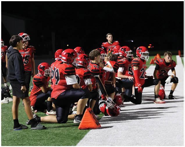 A group of people wearing red and black football uniforms are kneeling and sitting on the sidelines of a football field at night. Description generated by AI
