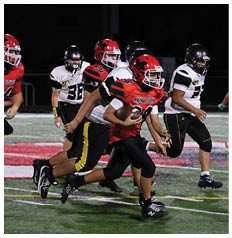 A group of people in football uniforms are actively running on a field at night with one person in a red jersey holding the ball and others in white and black jerseys chasing. Description generated by AI
