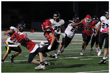 A group of people wearing football uniforms are actively playing on a field at night with one person in black and white being tackled by two people in red jerseys. Description generated by AI