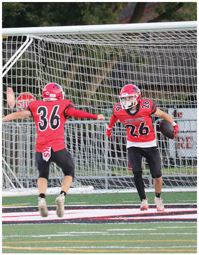 Two people in red football uniforms are celebrating near a goalpost on a grassy field with trees in the background. Description generated by AI