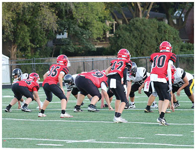 A group of people wearing red and black football uniforms are lined up on a green field facing a group in white and black uniforms, preparing for a play. Description generated by AI