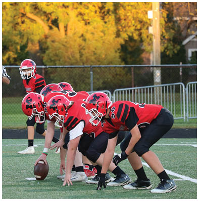 People in football uniforms are lined up on a field facing each other with a referee standing behind them and trees with autumn foliage in the background. Description generated by AI