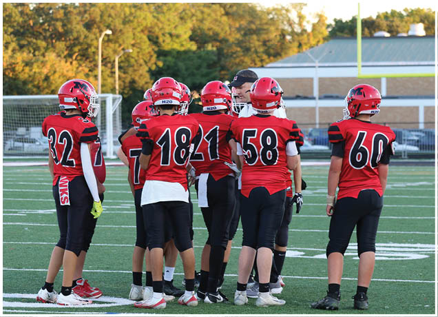 A group of people wearing red and black football uniforms and helmets are standing together on a football field during daylight. Description generated by AI