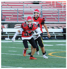 Three people wearing red and black football uniforms and helmets are running on a green field with empty bleachers in the background. Description generated by AI
