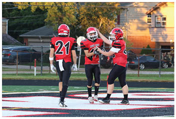 Three people wearing red and black football uniforms stand together on a sports field near the end zone with houses and trees in the background. Description generated by AI