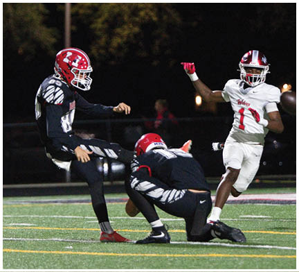 Two people in football uniforms are engaged in a play on a field with one person holding the ball on the ground while the other prepares to kick it, and a third person in a white uniform is running nearby. Description generated by AI