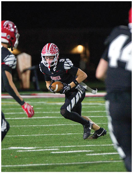 Three people in black football uniforms with red helmets are on a field at night, with one person holding a football and preparing to run. Description generated by AI