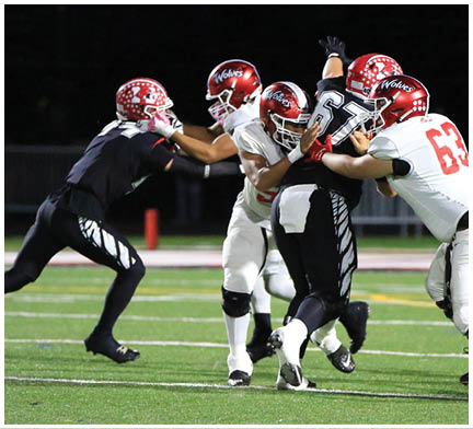 Four people in football uniforms are engaged in a play on a field under stadium lights, with one person in black attempting to block or tackle while three others in white and red helmets surround them. Description generated by AI