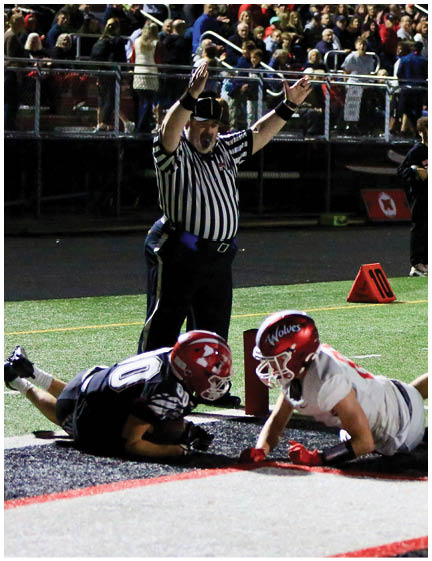 A person in a football uniform is diving toward the end zone while another person in a football uniform stands nearby and two people with cameras are capturing the moment on a football field with a crowd in the background. Description generated by AI