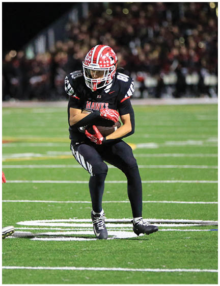 A person in a black uniform and red helmet is sliding on the grass field attempting to tackle another person in a white uniform with the number 88 running forward during a nighttime football game. Description generated by AI
