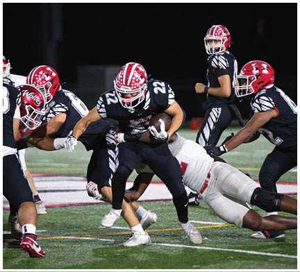 Two opposing American football teams in helmets and uniforms are lined up facing each other on a field with one player holding the ball ready to snap. Description generated by AI