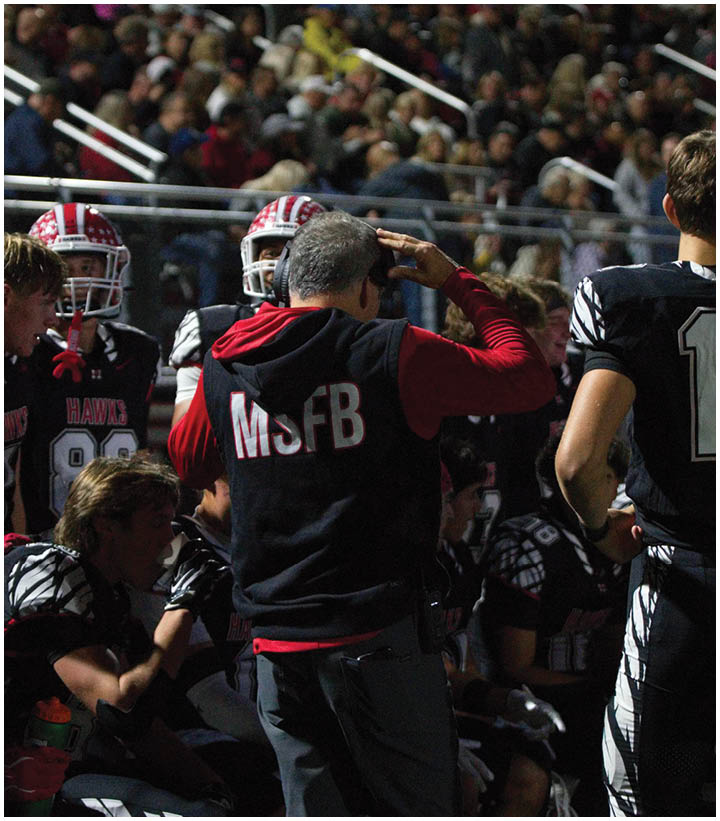 A person wearing a black vest with “MSFB" on the back is standing and saluting near a group of people in football uniforms on a field with a crowd in the background. Description generated by AI
