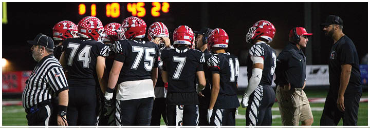 A group of football players in black uniforms and red helmets are gathered on a field with two referees and two coaches nearby under stadium lights. Description generated by AI