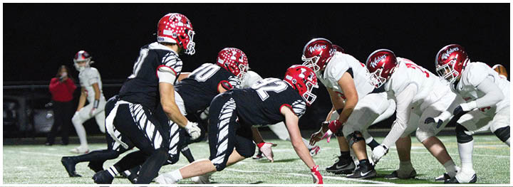 People in football uniforms are lined up on a field facing each other, preparing for a play during a nighttime game. Description generated by AI