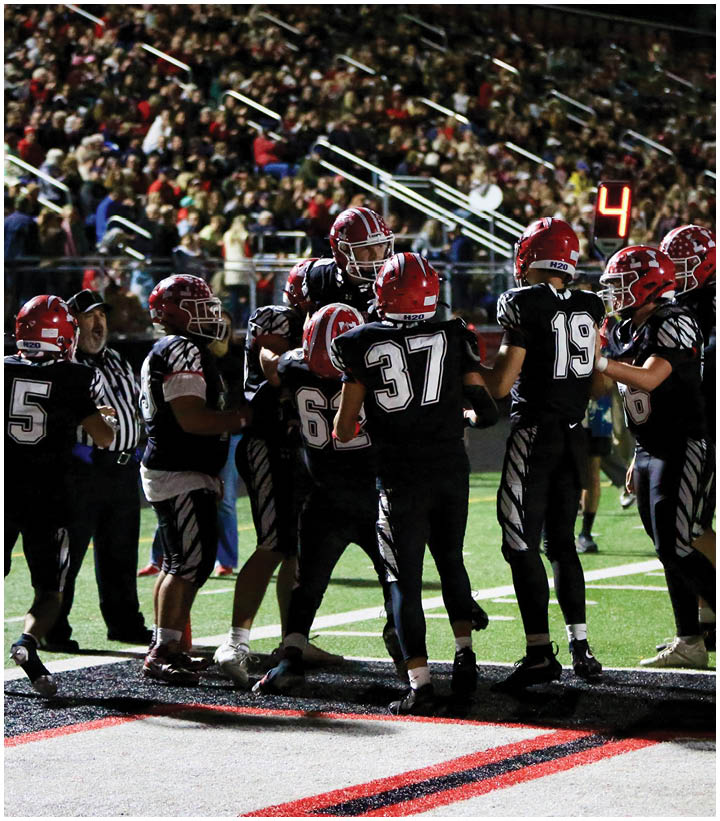 A group of football players wearing black uniforms and red helmets are gathered on a field near the sideline with a crowd visible in the background. Description generated by AI