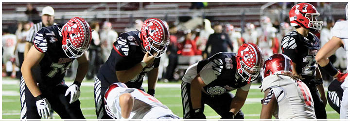 Players from two football teams are lined up on the field preparing for a play during a game in a stadium. Description generated by AI