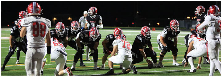 People in football uniforms are lined up on a field preparing for a play during a game at night. Description generated by AI