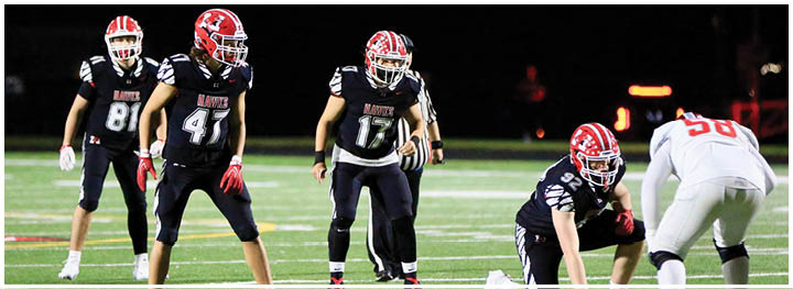 Four people in black football uniforms are positioned on a field facing one person in a white football uniform during a nighttime game. Description generated by AI