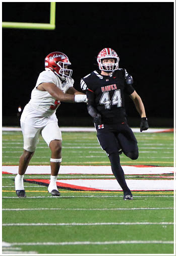Two people in football uniforms are actively playing on a field under stadium lights, with one person running and the other reaching out toward them. Description generated by AI