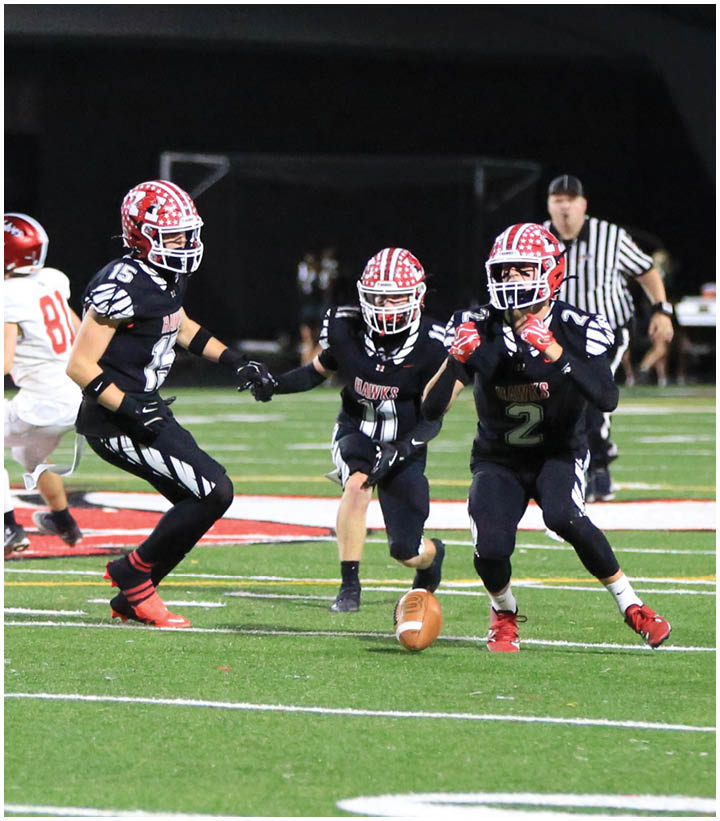 Three people in black football uniforms with red helmets are actively playing on a field with a football on the ground, while a referee and other players are visible in the background. Description generated by AI