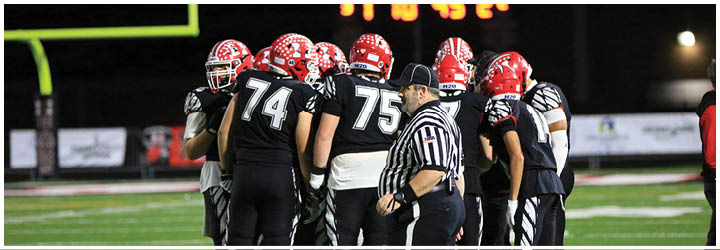 A group of football players wearing black uniforms and red helmets are huddled together on a field with a referee standing nearby and a scoreboard visible in the background. Description generated by AI