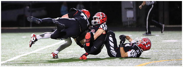 Three football players in black and red uniforms are engaged in a tackle on a field under stadium lights. Description generated by AI