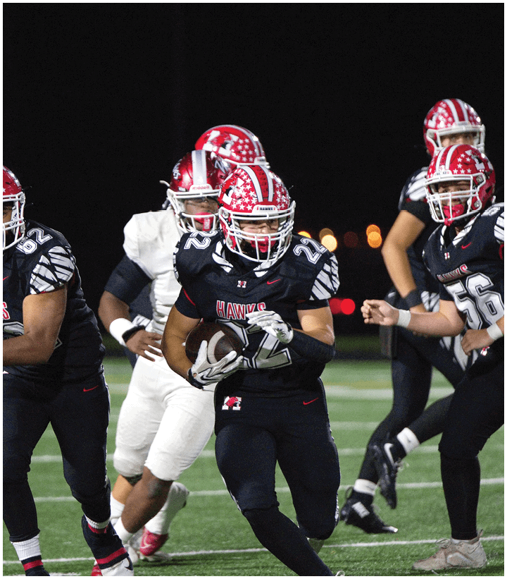 People wearing football uniforms are playing on a field at night with one person in black running while holding the ball and others in pursuit. Description generated by AI