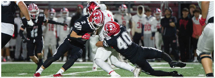 Two people in dark sports uniforms tackle a person in a white sports uniform during a football game with spectators in the background. Description generated by AI