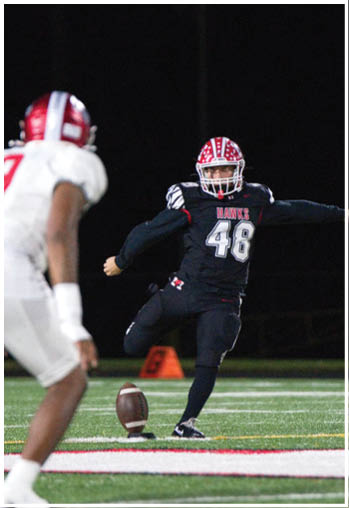 A person wearing a red and white helmet and a black football uniform with the number 48 is kicking a football on a field at night. Description generated by AI