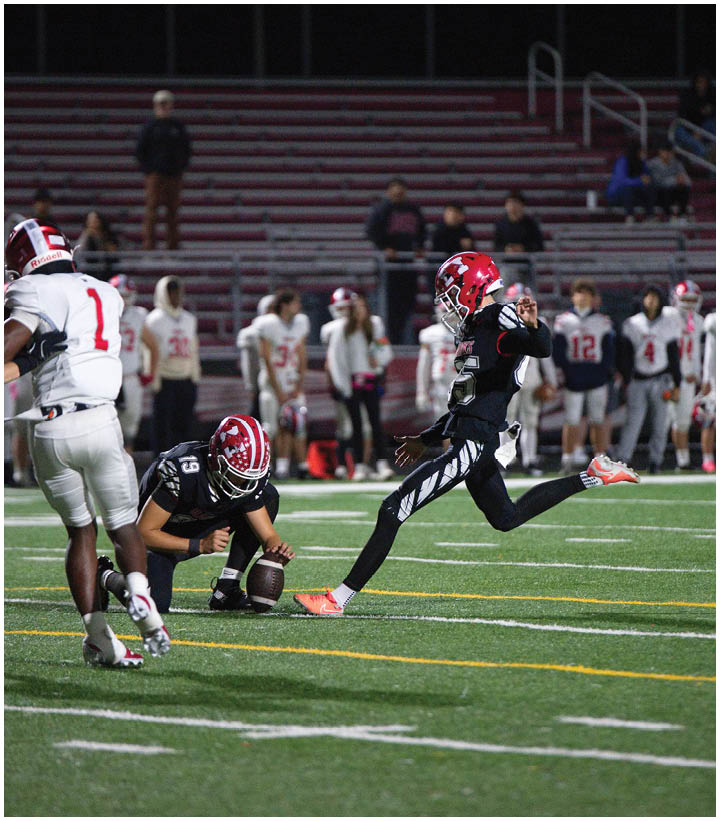 A person in a black sports uniform and red helmet is kicking a football held by another person on a green field with white and yellow lines, while a group of people in white uniforms watch from the background. Description generated by AI