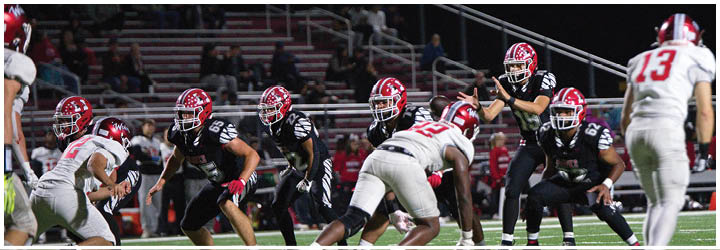 People in football uniforms are positioned on a field under stadium lights, preparing for a play during a nighttime game. Description generated by AI