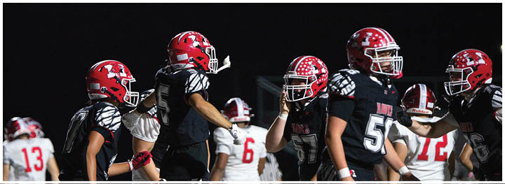 A group of football players wearing red helmets and black uniforms are gathered on a field at night. Description generated by AI