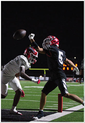 Two football players in helmets and uniforms compete near the end zone under stadium lights at night, with one player reaching to catch or block the ball. Description generated by AI