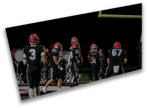 A group of people wearing black sports uniforms and red helmets are gathered on a football field near a goalpost at night. Description generated by AI