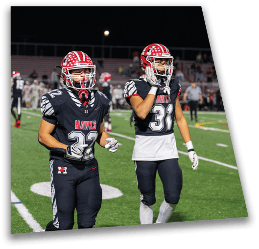Two people in football uniforms with red helmets stand on a green field during a nighttime game. Description generated by AI