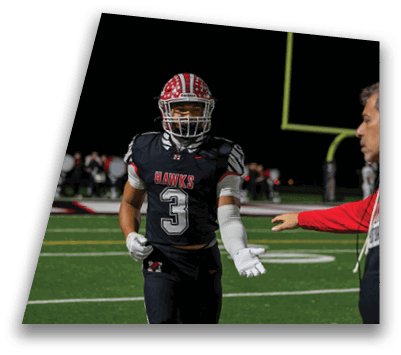 A person in a football uniform with the number 3 and the word “HAWKS" on the jersey is walking on a field at night while another person in a red and black jacket reaches out toward them. Description generated by AI