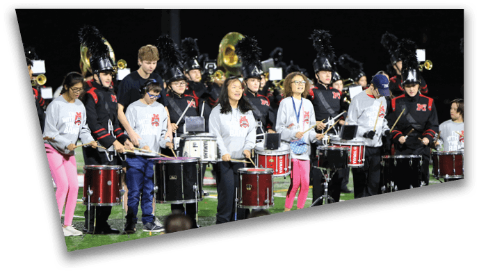 A group of people wearing band uniforms and casual clothing are playing drums together on a field at night. Description generated by AI