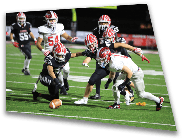Several people in football uniforms are reaching for a loose football on a green field during a nighttime game. Description generated by AI