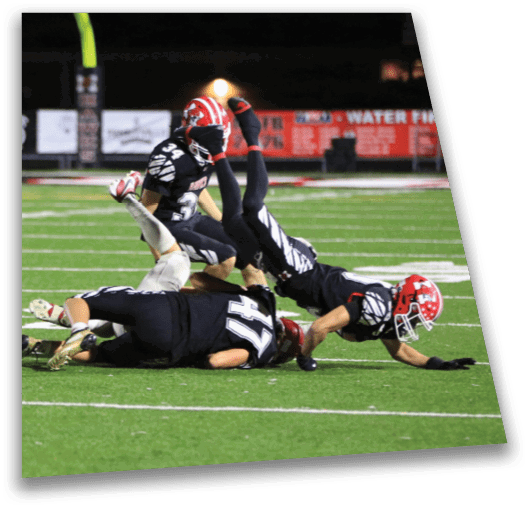 Three football players in black uniforms and red helmets are on the field with one player airborne and the others on the ground during a nighttime game. Description generated by AI