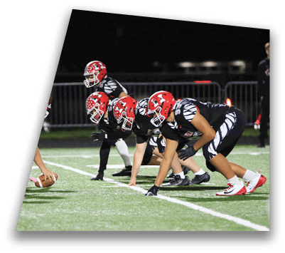 Two opposing football teams in red and white helmets are lined up facing each other on a field, ready for the snap. Description generated by AI
