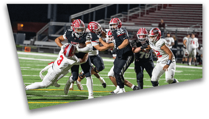 People wearing football uniforms are actively engaged in a play on a well-lit field with bleachers in the background. Description generated by AI