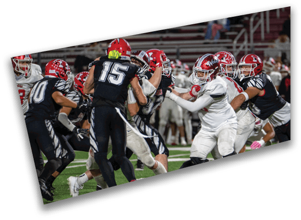 People wearing football uniforms are engaged in a play on a field under stadium lights with some players in black uniforms and others in white uniforms. Description generated by AI